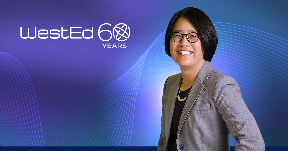 Photo of Jannelle Kubinec smiling with her hands in her pockets and wearing a grey blazer and black rimmed glasses.